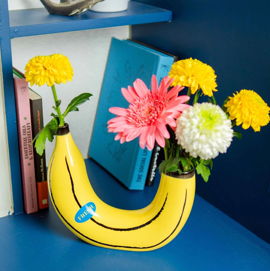 Banana-shaped vase with flowers on a blue shelf against a blue wall.