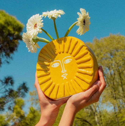 Yellow sun-shaped vase with floral design held against a blue sky and green trees